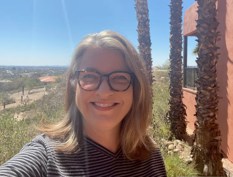 Head and shoulders image of Susan Miller-Cochran, smiling at the camera, with the Arizona desert in the background and the sun shining over her right shoulder